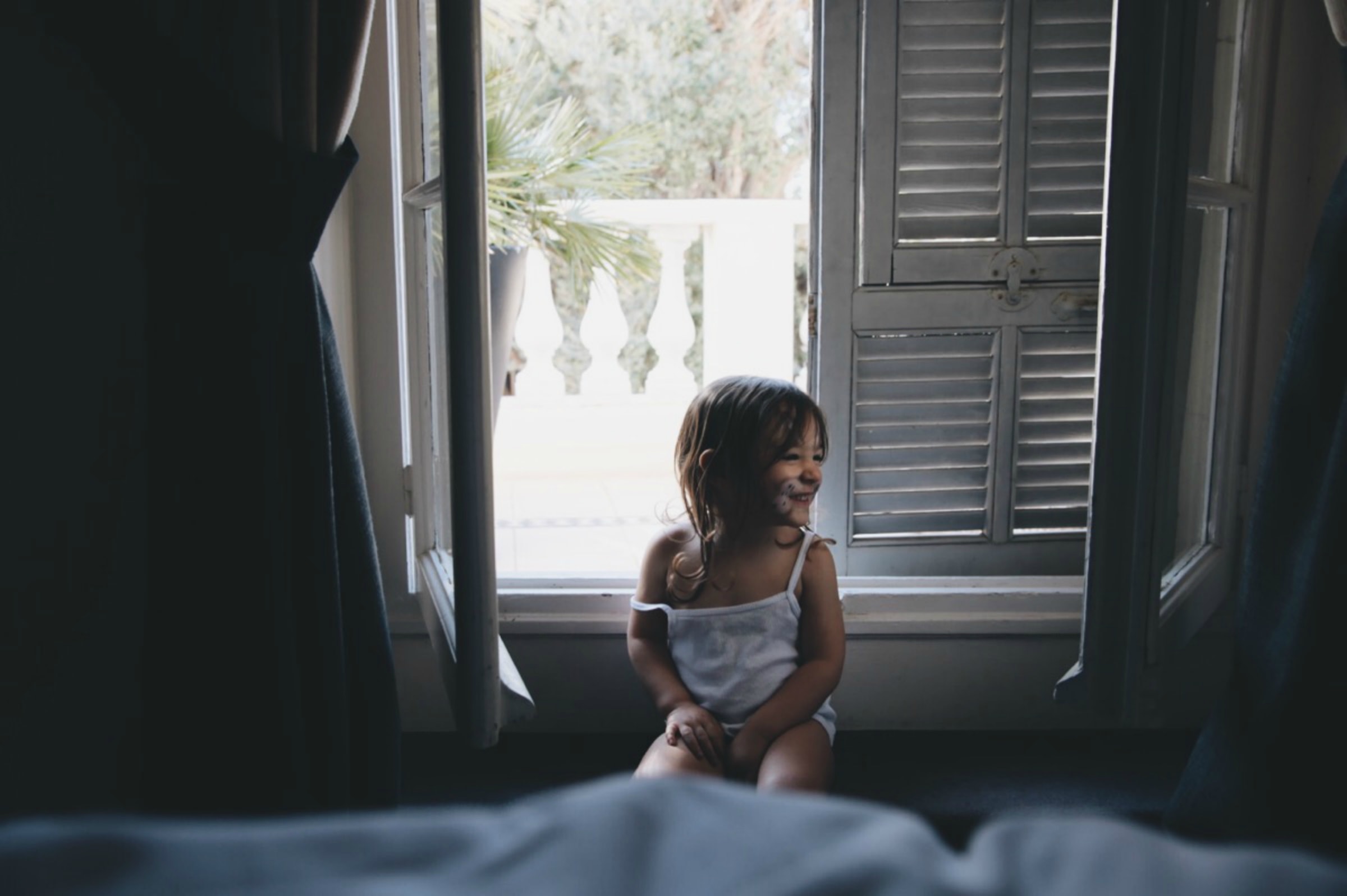 A smiling toddler standing at an open window in the morning