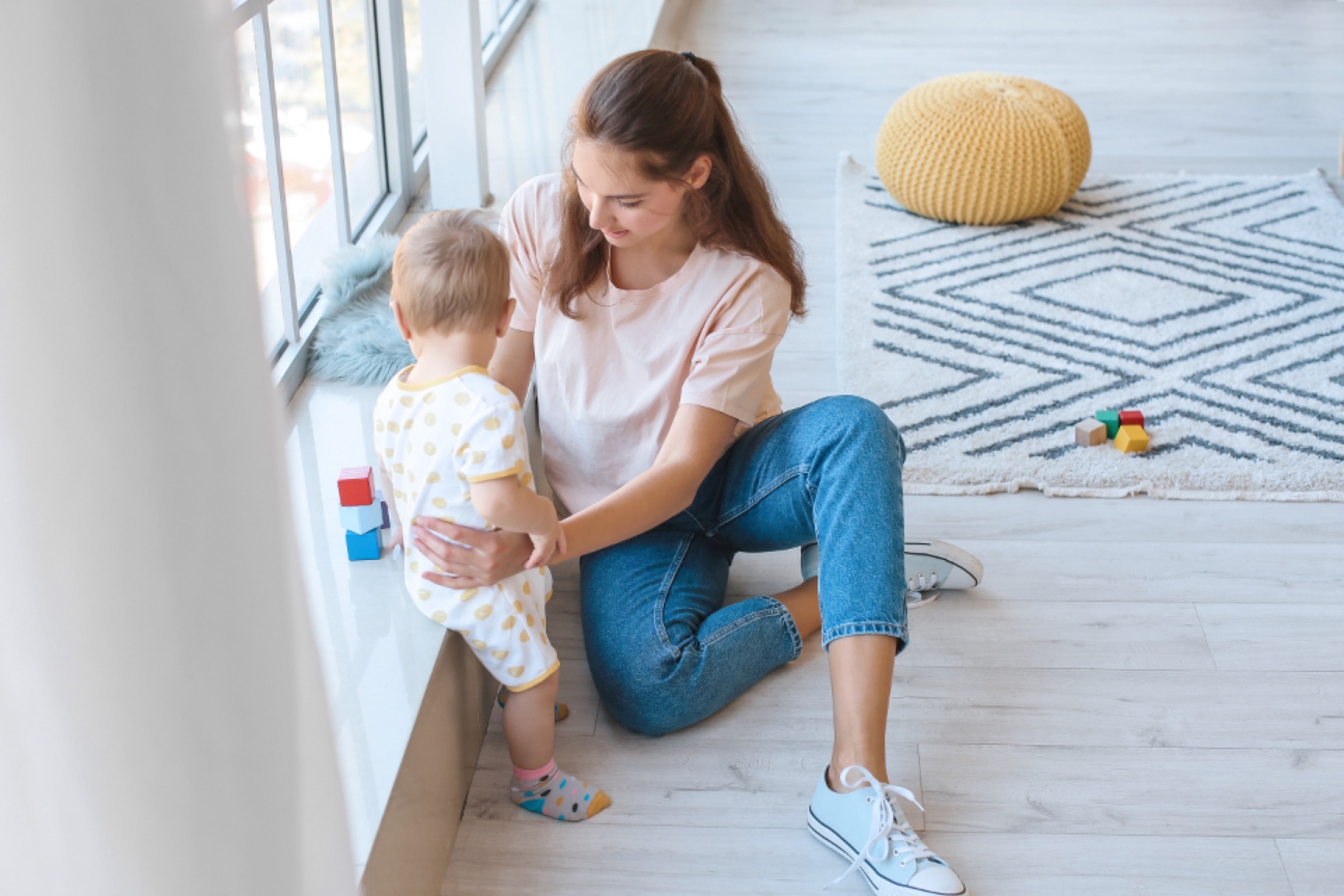 A mother sitting on the floor by a window playing with her baby