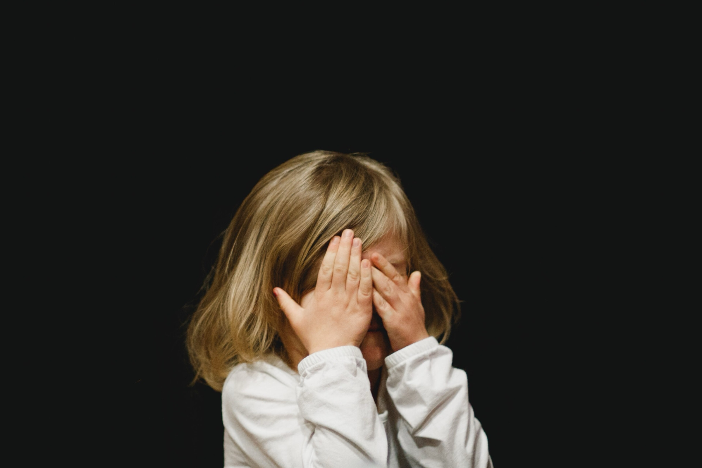 A young child with blonde hair covering her face with both hands against a dark background
