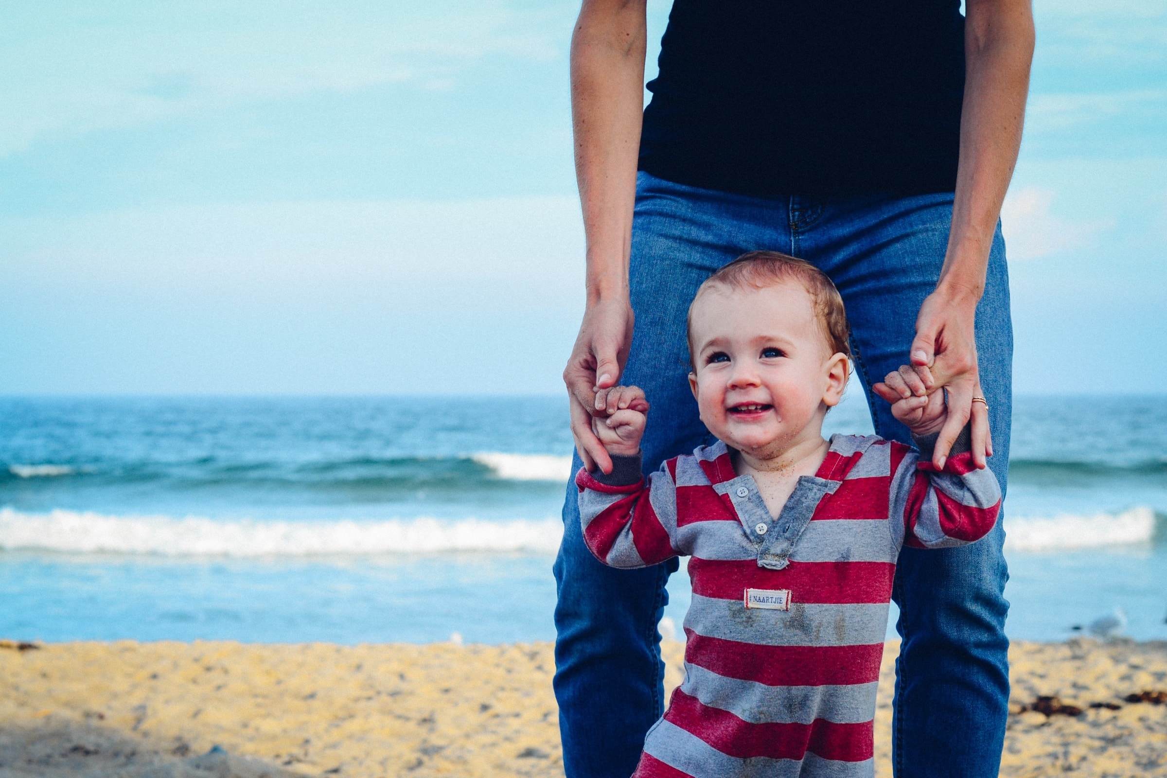 A toddler taking steps on the beach while holding a parent's hands